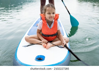 Close Up Of Cute Caucasian Girl In  Swim Life Vest Swimming And Her Father  On SUP Board. Family Paddleboarding On The Lake On Summer Day. Active Leisure With Kids. Family Local Getaway Concept