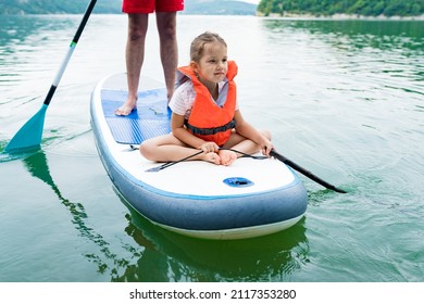 Close Up Of Cute Caucasian Girl In Swim Life Vest Swimming And Her Father On SUP Board. Family Paddleboarding On The Lake On Summer Day. Active Leisure With Kids. Family Local Getaway Concept