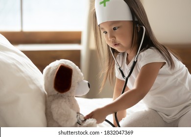 Close Up Cute Curious Asian Little Girl Wearing Doctor Uniform Using Stethoscope, Sitting On Cozy Bed, Pretty Toddler Child Kid Checking Listening To Toy Heart Beat, Pretending Pediatrician