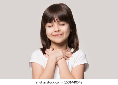 Close Up Of Cute Happy Small Girl Isolated On Grey Studio Background Hold Hands At Heart Chest Feel Grateful, Smiling Little Child With Eyes Closed Pray Thanking God High Powers, Faith Concept