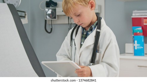 Close Up Of A Cute Little Boy Dressed Up As A Doctor In An Oversized Coat Using A Tablet Computer In A Medical Exam Room