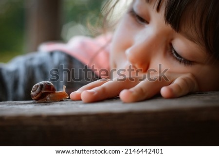 Close up of cute little girl intently watching small snail crawling along wooden bench while sitting comfortably in garden outdoor while, kids spending time in nature, child exploring environment