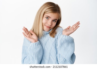 Close Up Of Cute Little Girl Shrugging Shoulders And Smiling Cute, Looking Innocent And Clueless, Standing In Blue Winter Sweater Against White Background