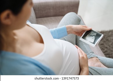 Close up of cute pregnant belly and x ray ultrasound scan of baby. asian young pregnant female relax on couch sofa looking at her child picture in hands sweet. motherhood concept third trimester