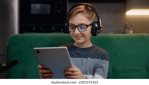 Close up of cute smiling smart boy using digital tablet browsing, streaming and watching videos online with headphones at home. Beautiful young guy holding tablet computer sitting on sofa at home in - Powered by Shutterstock