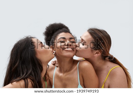 Close up of cute young women in colorful underwear kissing their friend while posing together isolated over white background. Friendship, beauty, body positive concept. Horizontal shot