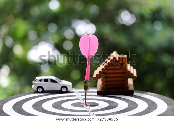 close up dart arrow hitting on target center of\
dartboard with toy car and wood house background, success and\
business concept