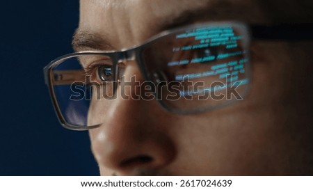 Close up of data scientist face is closely examines data. The glow of code lines reflecting in software engineer glasses and eyes.