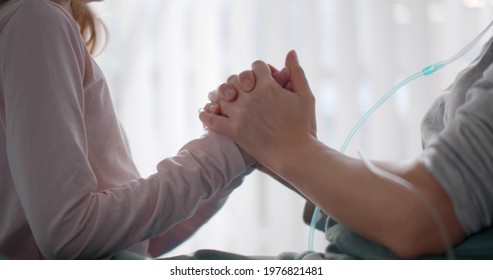 Close Up Of Daughter Supporting Ill Mother Lying On Hospital Bed. Sick Woman Holding Hands With Little Daughter Visiting In Hospital Ward. Family And Healthcare Concept