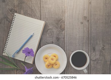 A Close Up Of A Desktop With Blank Notepad, Cup Of Coffe And Donuts On It. Wooden Background, Top View