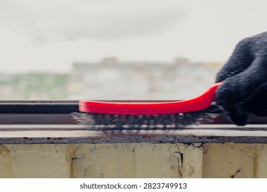 Close detail of gloved hand using red wire brush to clean metal balcony frame before painting, removing rust, corrosion, and old paint from narrow sill with blurred city buildings behind