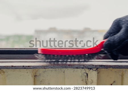 Close detail of gloved hand using red wire brush to clean metal balcony frame before painting, removing rust, corrosion, and old paint from narrow sill with blurred city buildings behind