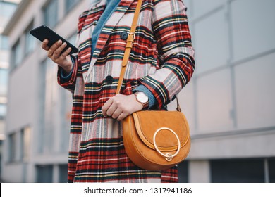 Close Up Detail Of Modern Day Female Accessories - Stylish Young Woman Wearing An Overcoat With A Chequered Pattern And A Wrist Watch While Holding A Fancy Camel Bag And Typing On Her Mobile Phone.