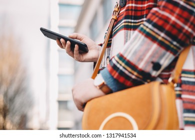 Close Up Detail Of Modern Day Female Accessories - Stylish Young Woman Wearing An Overcoat With A Tartan Pattern And A Wrist Watch While Holding A Fancy Yellow Bag And Typing On Her Mobile Phone.