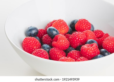 A Close Up Detail Of Superfood Mixed Berries Of Raspberries And Blueberries In A White Bowl On A Plain White Surface And Background.