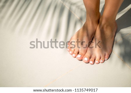 Close up detail view of bare tanned female feet with white pearl pedicure on white sand with a palm tree shadow. Travel and holiday lifestyle.