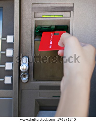 Close up detail view of a young woman hand holding and inserting a red credit card in a cash point machine with a reflective screen, outdoors. Finances and money availability funds.