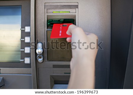Close up detail view of a young woman hand holding and inserting a red credit card in a cash point machine with a reflective screen, outdoors. Finances and money availability funds.