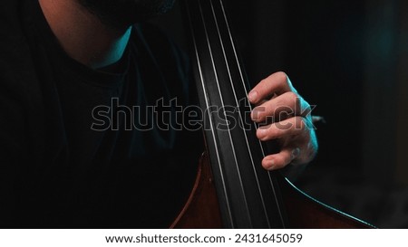 Close up details of the fingers of a musician's hand on the fingerboard of his double bass on a dark background with blue backlight