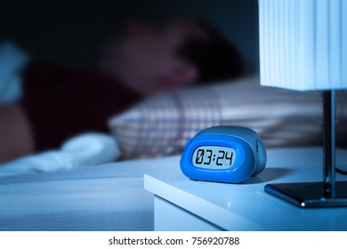 Close Up To Digital Alarm Clock On Nightstand In Bedroom. Man Sleeping In Bed In The Background. Dreamy Blur Effect.