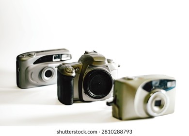 Close up of a digital DSLR camera in sharp focus surrounded by two blurred vintage point-and-shoot film cameras, isolated on a bright white background.
