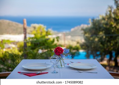 Close Up Dinner Table On The Outside With A Sea View, Red Flower And Plates On Table