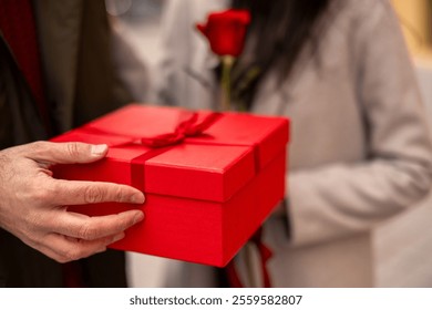 Close up as diverse couple exchanges gifts, featuring a man's hand holding a red present. They stand outdoors, creating a romantic professional setting for Valentine’s Day celebration. - Powered by Shutterstock