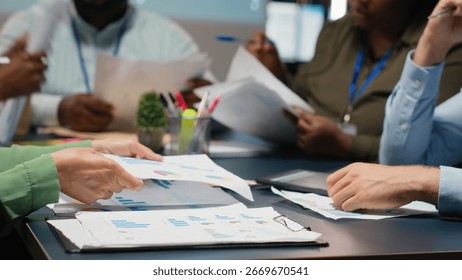Close up of diverse specialists evaluating data numbers on archived files, exchanging reports and insights to establish clear enterprise objectives in strategy room late at night. Camera B.