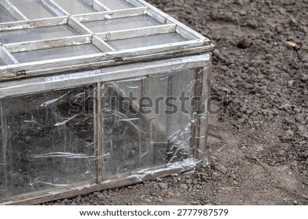 Close up of a DIY cold frame box constructed with vintage wood and plastic film, resting on plowed soil to provide warmth and shelter for early spring plants in a sustainable home garden
