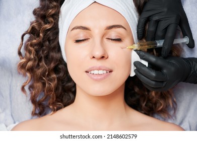 Close Up Of Doctor Beautician Hands In Sterile Gloves Making Injection Into Young Woman Face. Female Client Wearing White Headband While Receiving Facial Treatment At Beauty Salon.