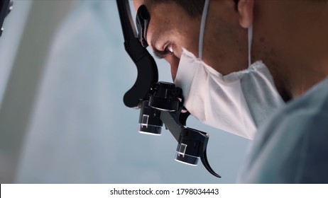 Close Up Of Doctor Face With Surgical Loupe, Sterile Mask And Protective Hair Cover Looking Down. Action. Male Surgeons Looking Very Serious And Concentrated On Their Work.