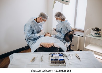 Close Up Of Doctor Hand Holding Forceps With Tampon While Surgeon And Assistant Performing Plastic Surgery On Blurred Background. Medical Team Using Various Surgical Instruments In Operating Room.