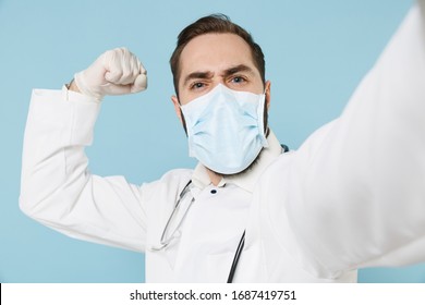 Close Up Doctor Man In Medical Gown Face Mask Gloves Isolated On Blue Background. Epidemic Pandemic Coronavirus 2019-ncov Sars Covid-19 Flu Virus. Doing Selfie On Mobile Phone Showing Biceps Muscles