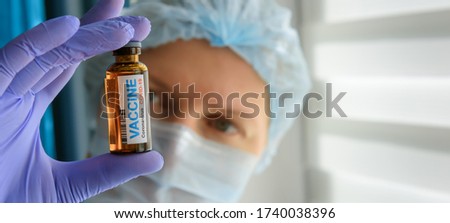 Close up doctor in medical mask and latex gloves holds in her hand vial with covid-19 vaccine. Nurse in white coat  looks at the bottle with coronavirus vaccine, blurred background.