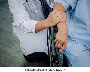 Close Up Doctor's Hands Holding Elderly Patient 's Hand In A Wheelchair. Male Doctor In White Suit Visit And Encourage Senior Woman Patient Dressed In Light Blue In Hospital. Elderly Care Concept.