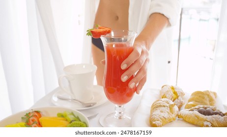 CLOSE UP DOF: Young Caucasian woman in white shirt picking up freshly squeezed fruit juice for breakfast in stunning luxury hotel room and going to the terrace with view on sailboat harbor - Powered by Shutterstock