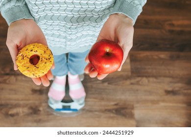 Close up donut and apple in unrecognizable hand of woman or girl standing on scales. Flour of choice. Diet. Sweet donut in hand, apple in hand, red apple, endless diet. Concept of beauty and health.