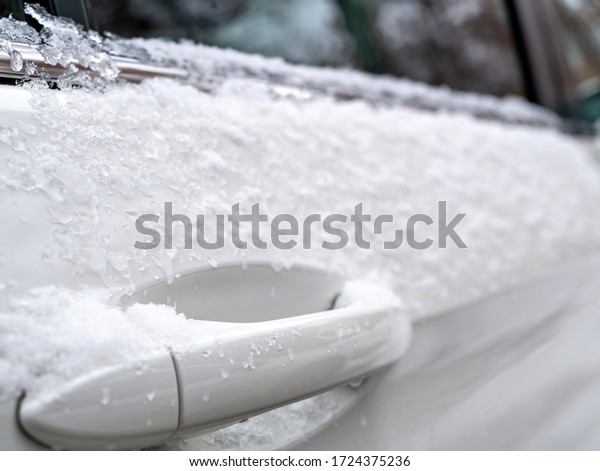 A close up of the door handle of a white car\
after snow has fallen on the\
vehicle.