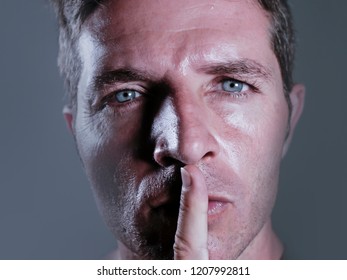 close up dramatic studio portrait of young handsome and attractive man with finger on his closed mouth warning to shut up and not to speak in censorship and freedom of speech coercion