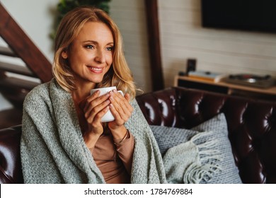 Close up of dreaming middle aged woman sitting in living room with cup of coffee or tea enjoying under blanket