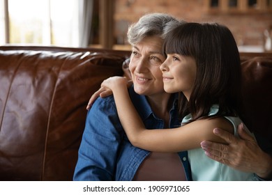 Close up dreamy mature woman and little girl hugging, looking to aside, sitting on couch at home, older grandmother and adorable granddaughter dreaming about good future, planning, two generations