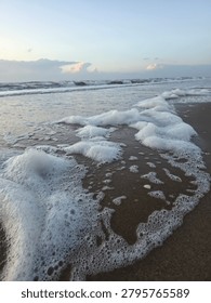 A close up dynamic shot of thick white sea foam washing over wet sandy beach with ocean waves in the background