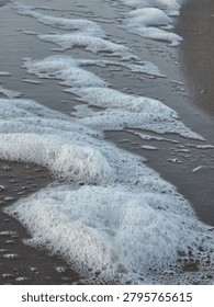 A close up dynamic shot of thick white sea foam washing over wet sandy beach with ocean waves in the background