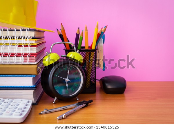 Close up\
educational materials on wooden desk, construction helmet, stack of\
book, calculator, pencils, clock, dividers, stapler and  computer\
mouse isolated pink\
background.