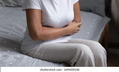 Close Up Elderly Female Arms Embracing Hurt Belly. Middle Aged Senior Unhealthy Woman Sitting On Bed, Suffering From Strong Stomach Ache, Feeling Morning Pain, Pancreatitis Gastritis Diarrhea Concept.