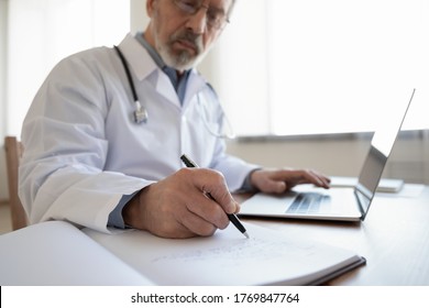 Close Up Of Elderly Grey-haired Male Doctor Or GP Sit At Desk In Hospital Write In Paper Medical Journal Work On Laptop, Serious Old Mature Physician Make Notes Fill Patient Anamnesis In Book Register