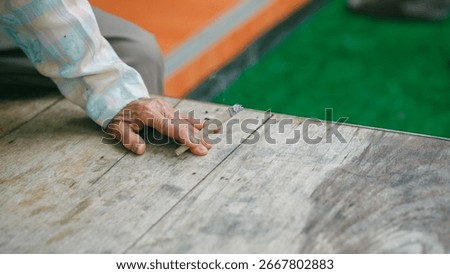Close up of elderly hand holding burning cigarette with smoke rising on wooden table, concept of smoking addiction, health risk, lung disease, and harmful lifestyle habits.