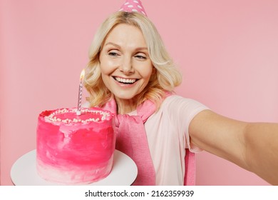 Close Up Elderly Happy Fun Woman 50s In T-shirt Birthday Hat Hold Cake With Candle Do Selfie Shot Pov On Mobile Phone Isolated On Plain Pastel Pink Background Studio. Celebration Party Holiday Concept