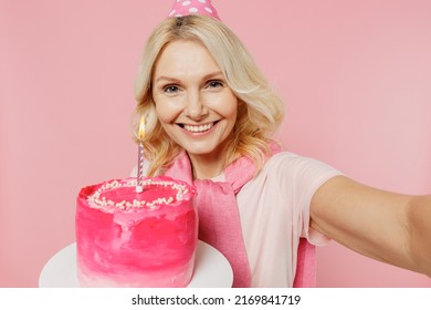 Close Up Elderly Happy Woman 50s Wear T-shirt Birthday Hat Hold Cake With Candle Do Selfie Shot Pov On Mobile Phone Isolated On Plain Pastel Pink Background Studio. Celebration Party Holiday Concept