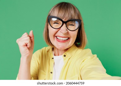 Close Up Elderly Smiling Happy Fun Woman 50s In Yellow Shirt Glasses Do Selfie Shot Pov On Mobile Phone Do Winner Gesture Celebrate Isolated On Plain Green Background Studio People Lifestyle Concept.